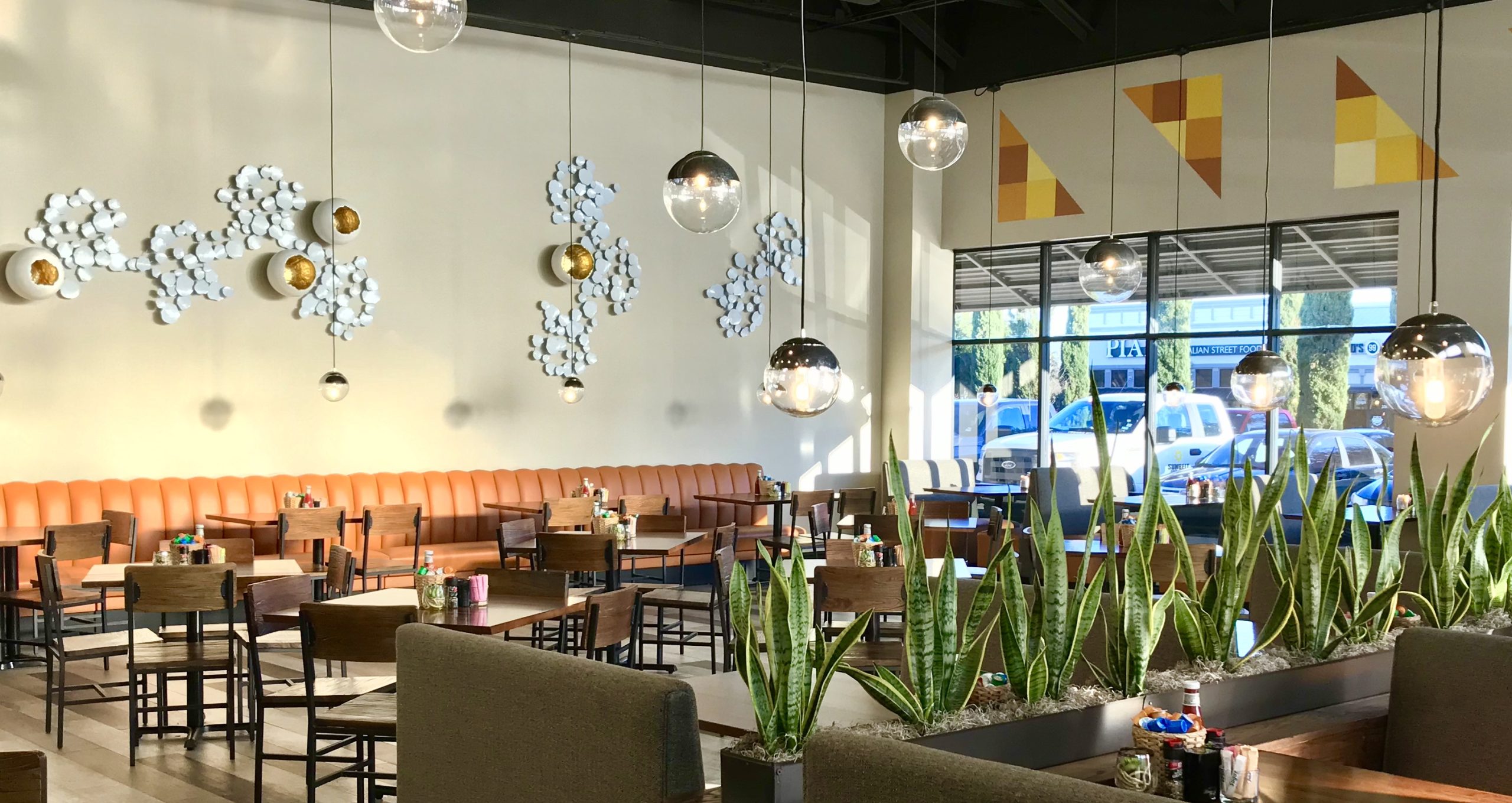 The inside of the restaurant "Biscuit" featuring orange booths, wooden tables and chairs, plants, sphere lights, white and gold decorative egg wall art and orange and yellow colored triangular wall art.