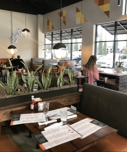 The inside of the restaurant "Biscuit" featuring plants, sphere lights, and orange and yellow colored triangular wall art. Menus and waters are on a table. A girl's back can be seen standing at the hostess stand.