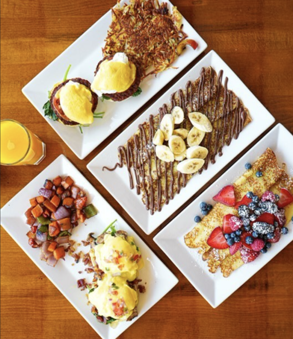 Four plates of food and a cup of orange juice on a table. The food includes eggs Benedicts, hash browns, sweet potato hash, banana Nutella crepes, and berry covered crepes.